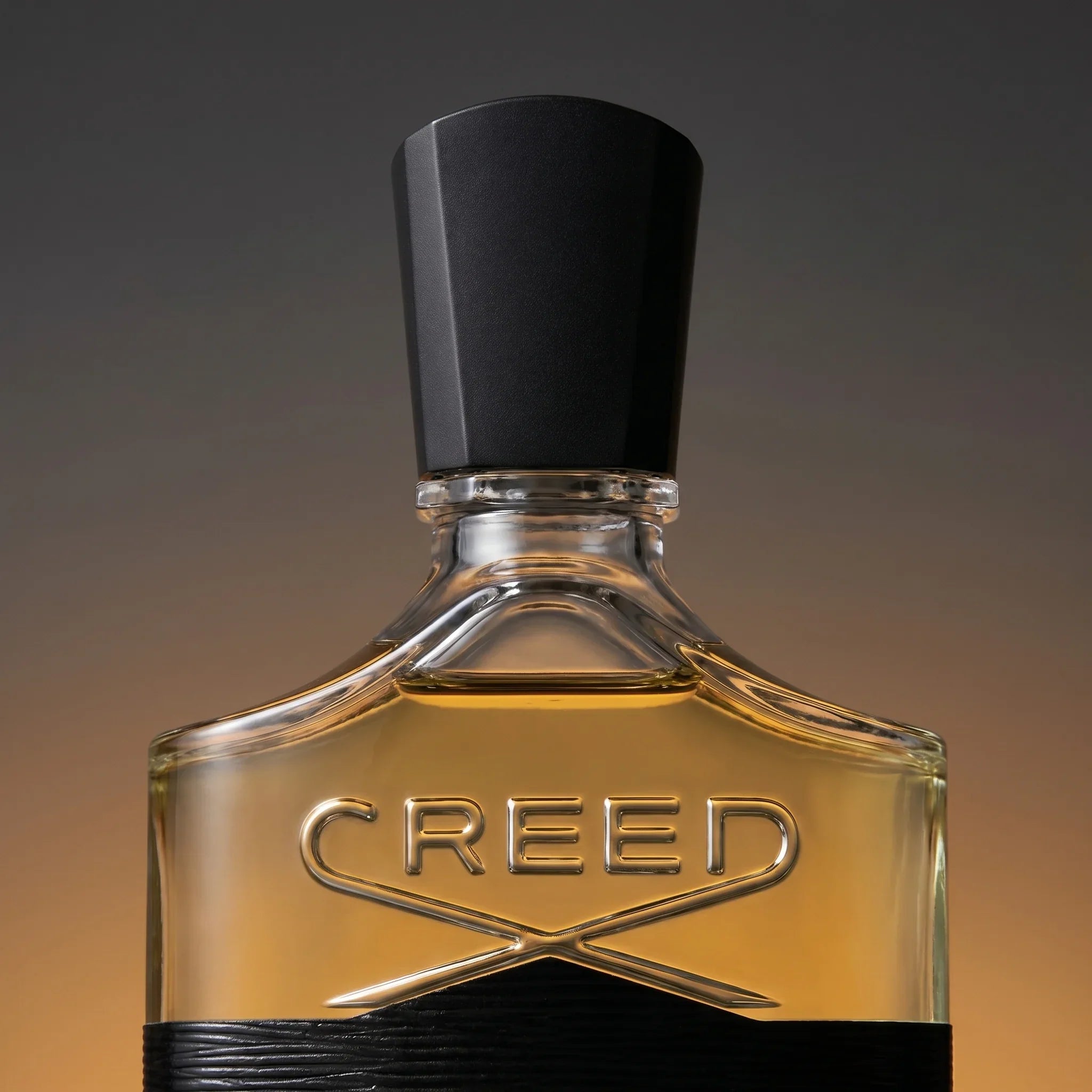 Macro detail of Creed Aventus cap and shoulders — black rectangular cap with silver collar and crossed-swords CREED engraving over golden-amber juice
