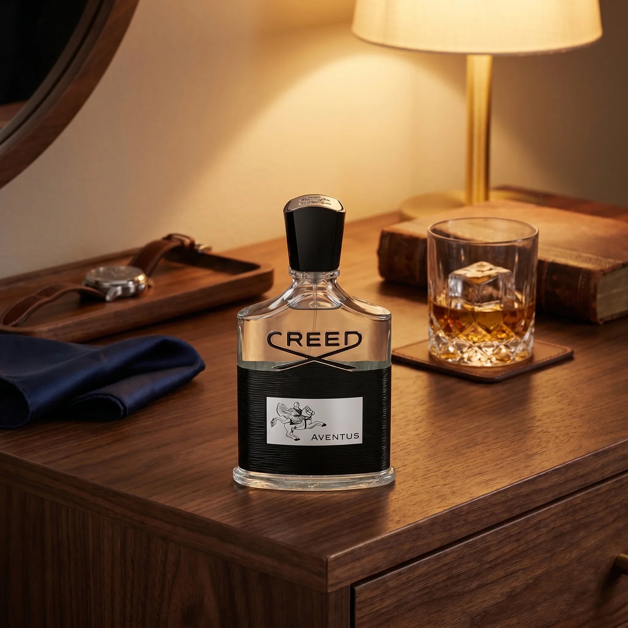 Creed Aventus on a dark walnut vanity with leather wristwatch, navy pocket square and whiskey glass in warm evening lamp light
