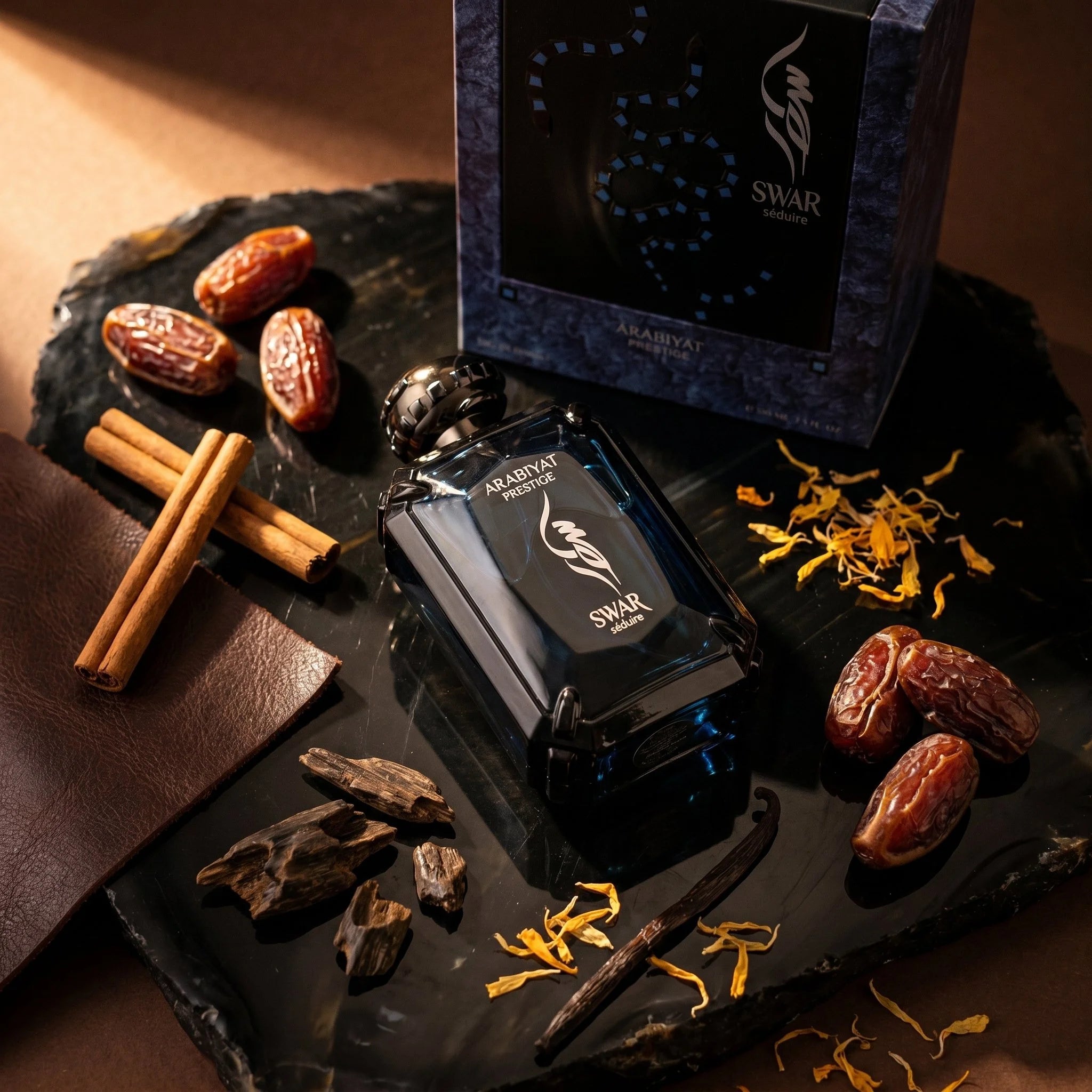 Arabiyat Prestige Swar Séduire flatlay with cinnamon, dates, marigold, oud, leather, and vanilla on dark slate