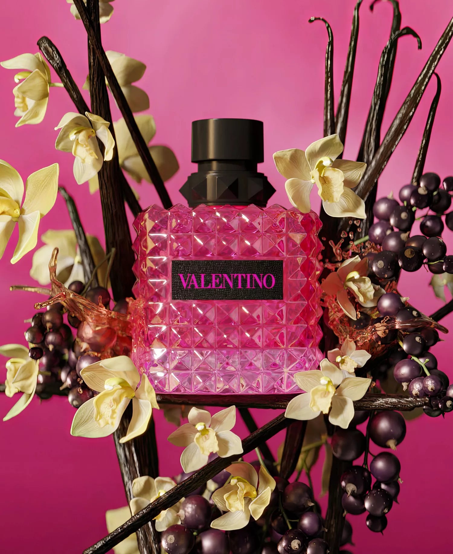 Pink perfume bottle with 'Valentino' branding surrounded by flowers and fruits on a pink background