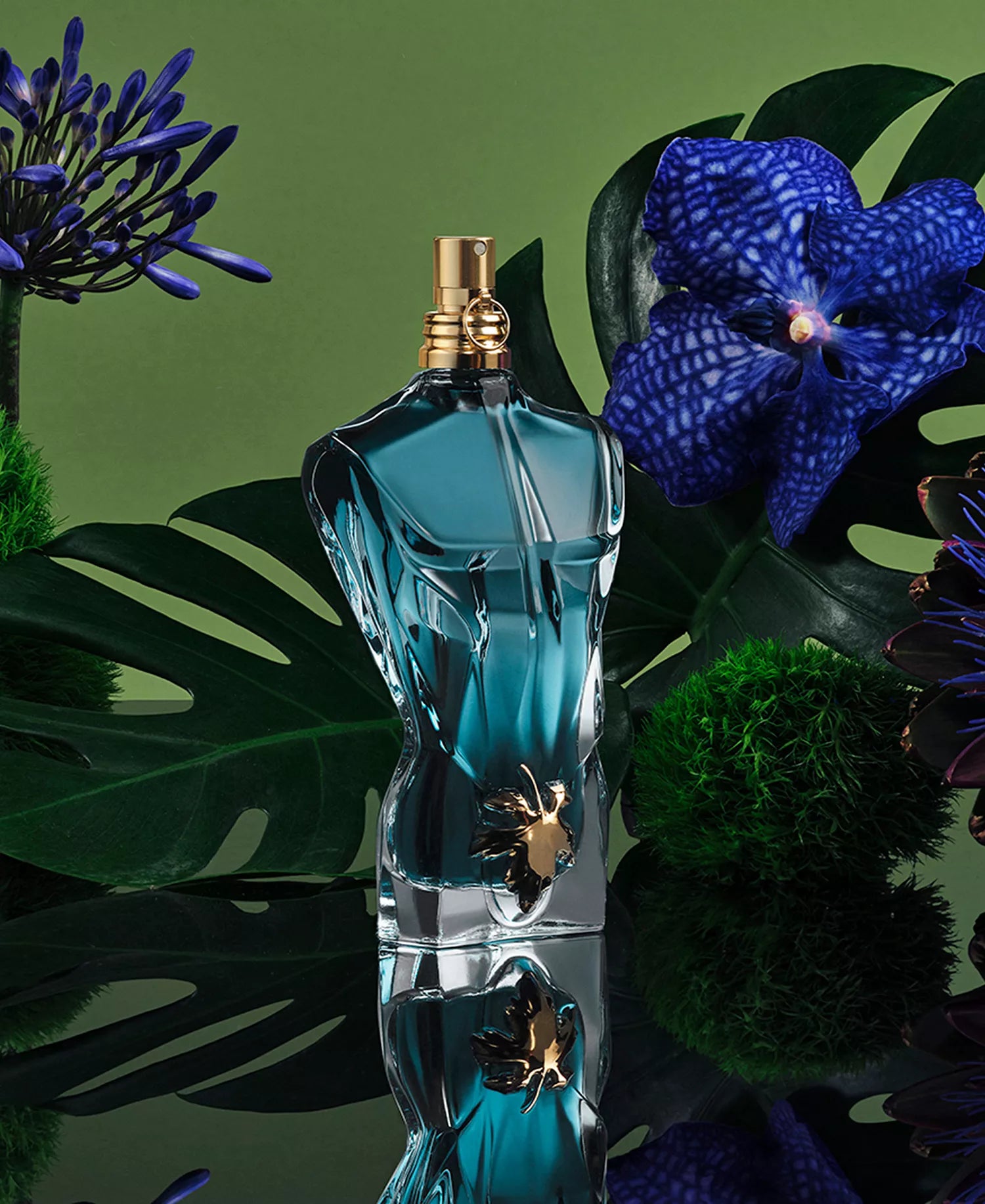 Blue perfume bottle with gold cap on a reflective surface surrounded by green and purple foliage.