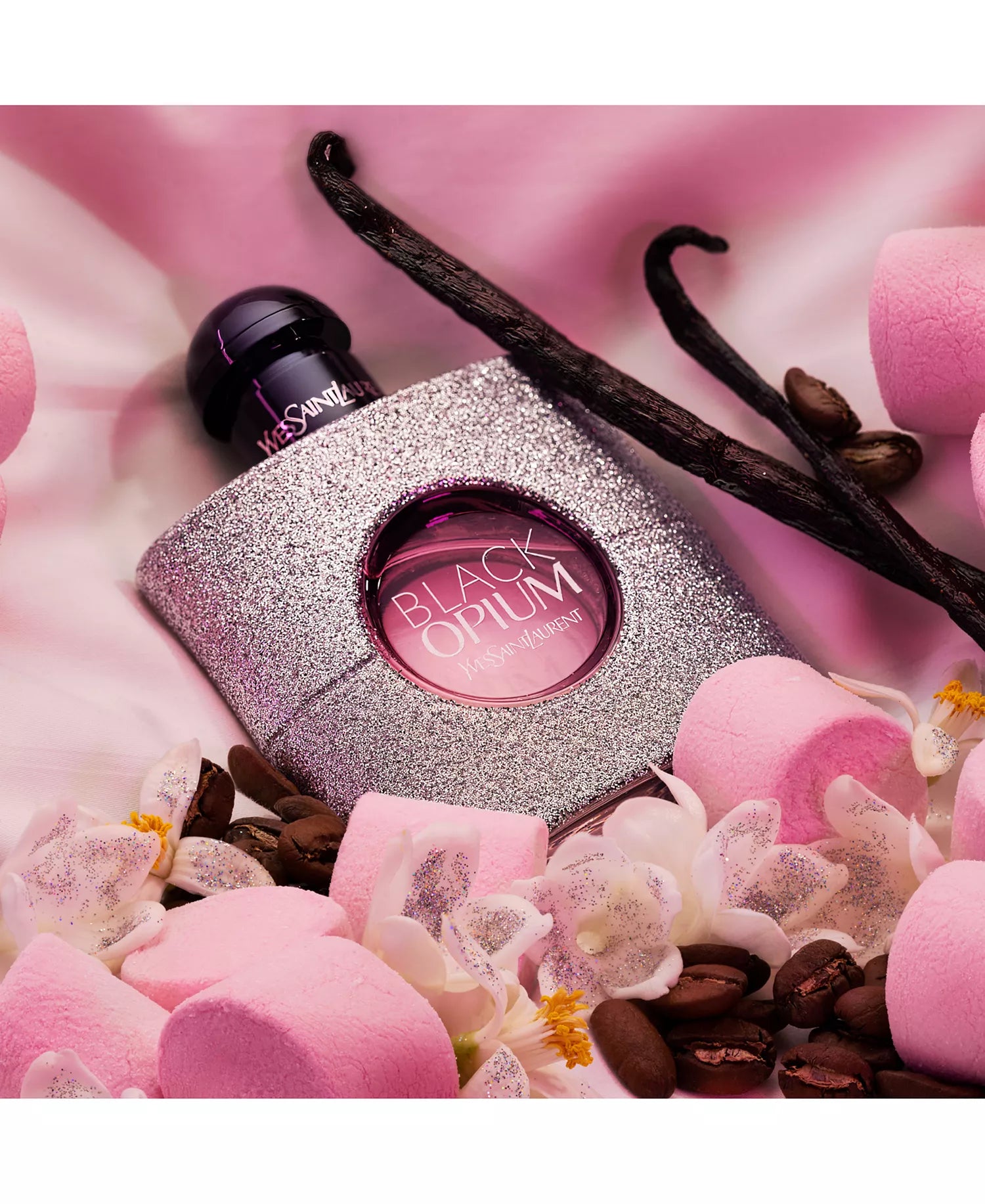 YSL Black Opium perfume bottle surrounded by vanilla beans, flowers, and pink petals on a pink background