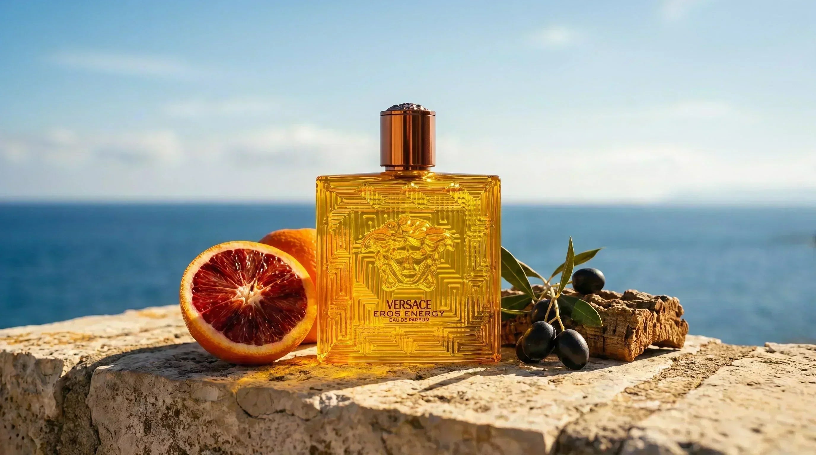 Versace Eros Energy EDP amber gold bottle on Mediterranean limestone wall with blood orange and sea backdrop