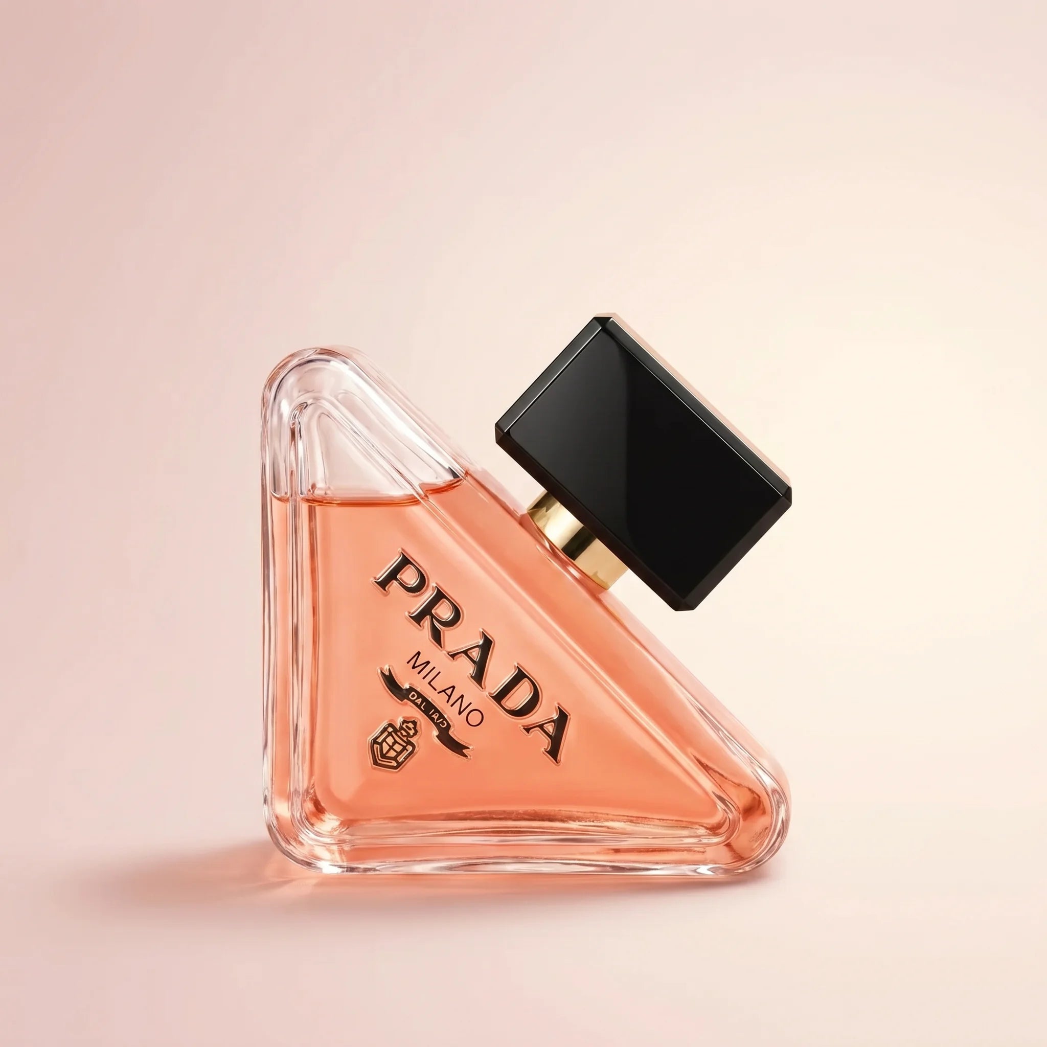 Prada Paradoxe Eau de Parfum 3 oz bottle — clear triangular glass with peach-coral juice and gold PRADA Milano logo on a soft blush backdrop