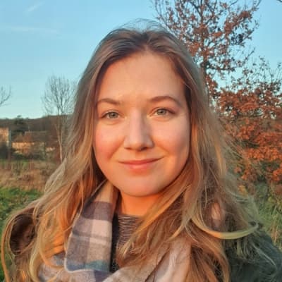 Woman with long blonde hair standing outdoors with trees and blue sky in the background