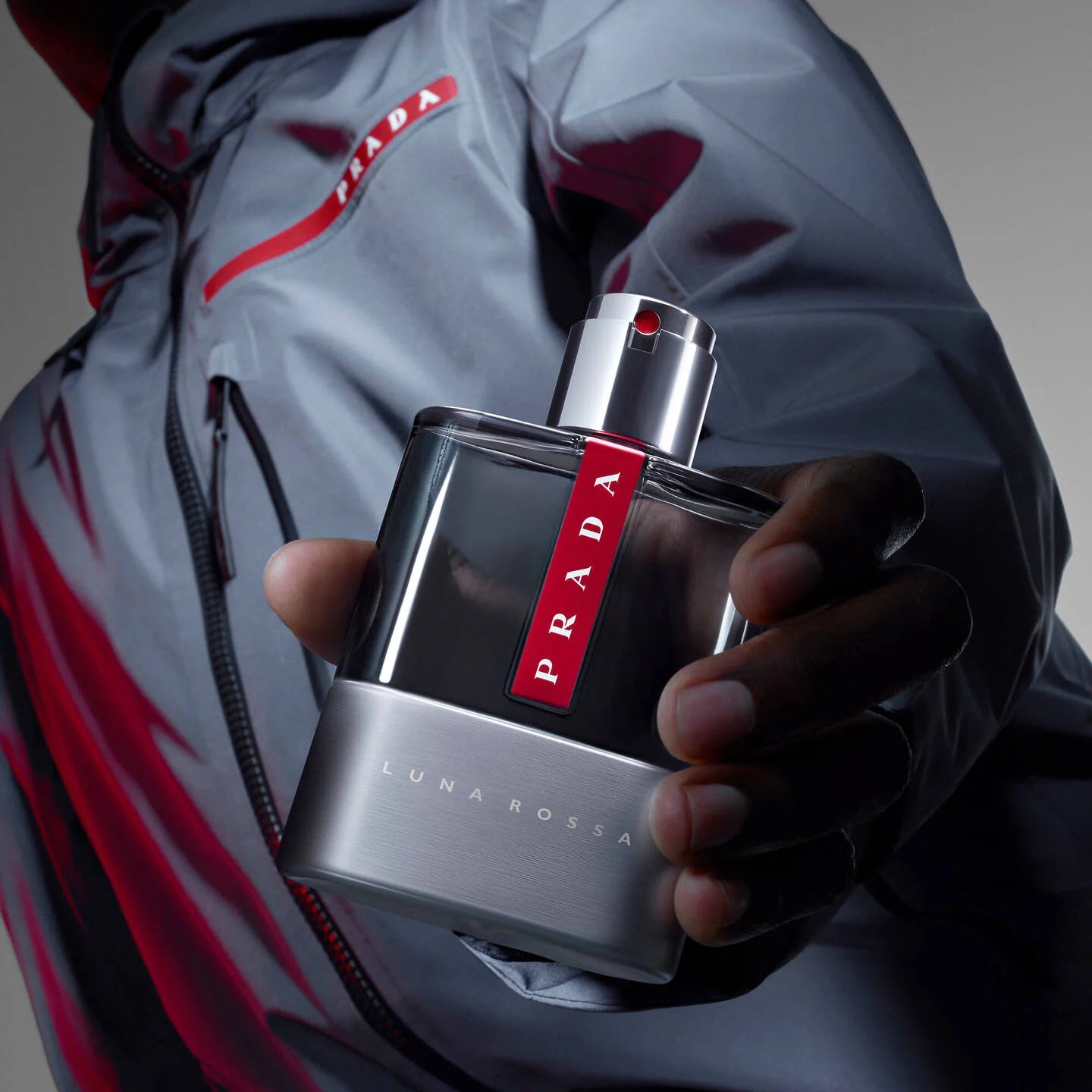 Person holding a Prada Luna Rossa perfume bottle with a gray and red jacket.