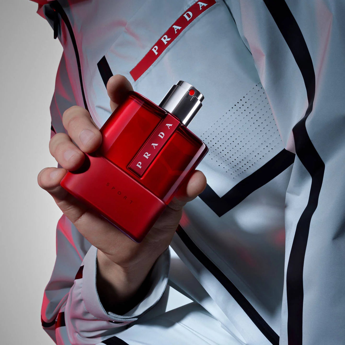 Red Prada perfume bottle held by a hand with a blurred Prada jacket in the background.