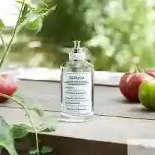 Replica From the Garden EDT by Maison Margiela placed on a wooden table with tomatoes and green leaves, reflecting its natural, unisex scent.