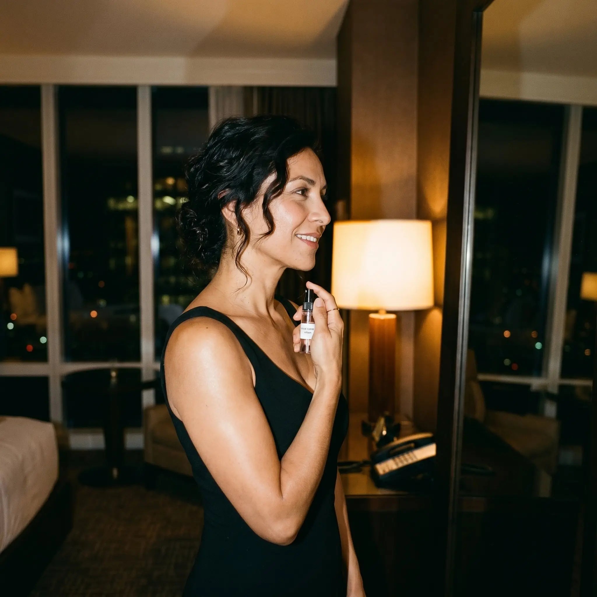 Woman in black dress applying Afnan 9PM Night Out decant in luxury hotel room with city view
