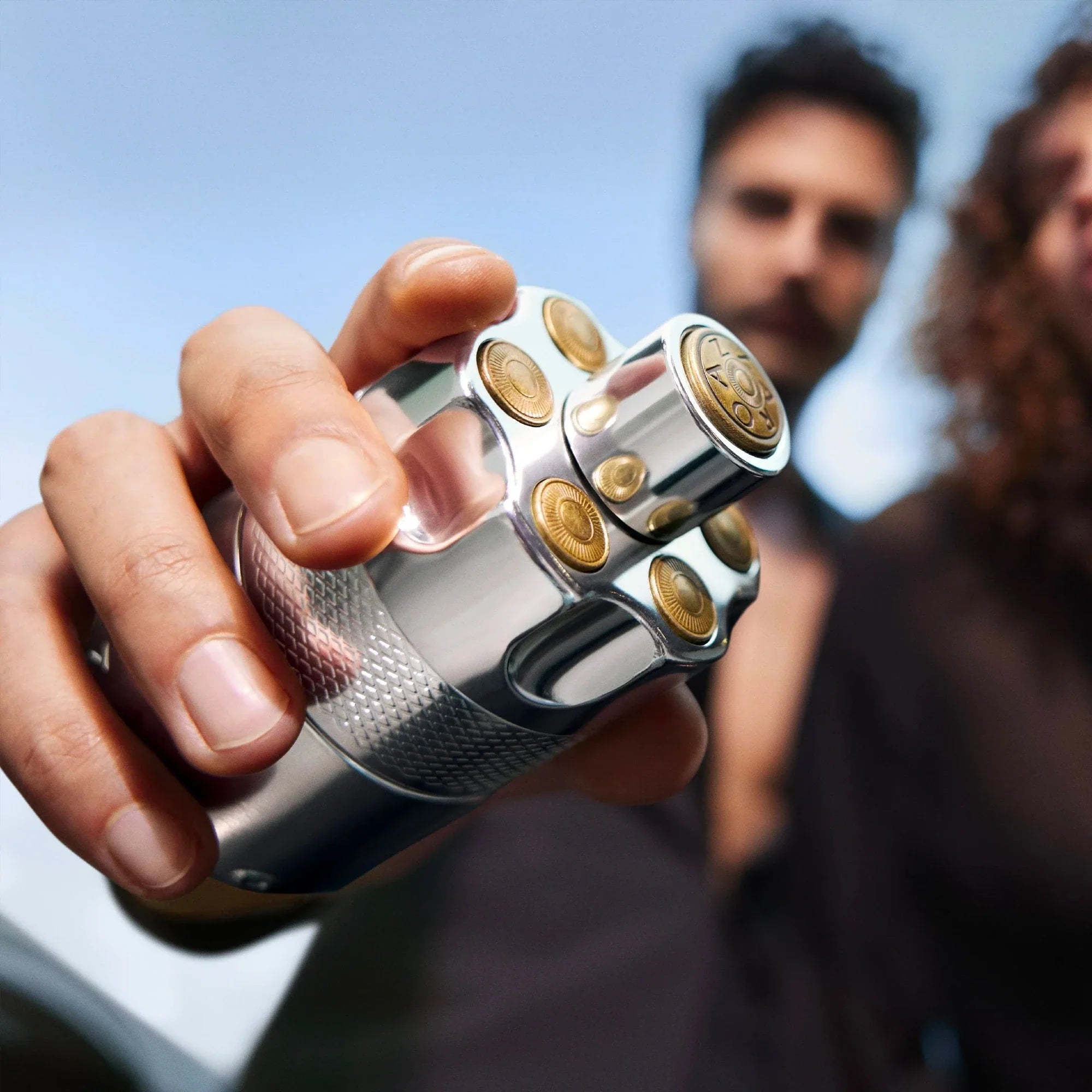 Hand holding a metallic object with gold accents against a blurred outdoor background