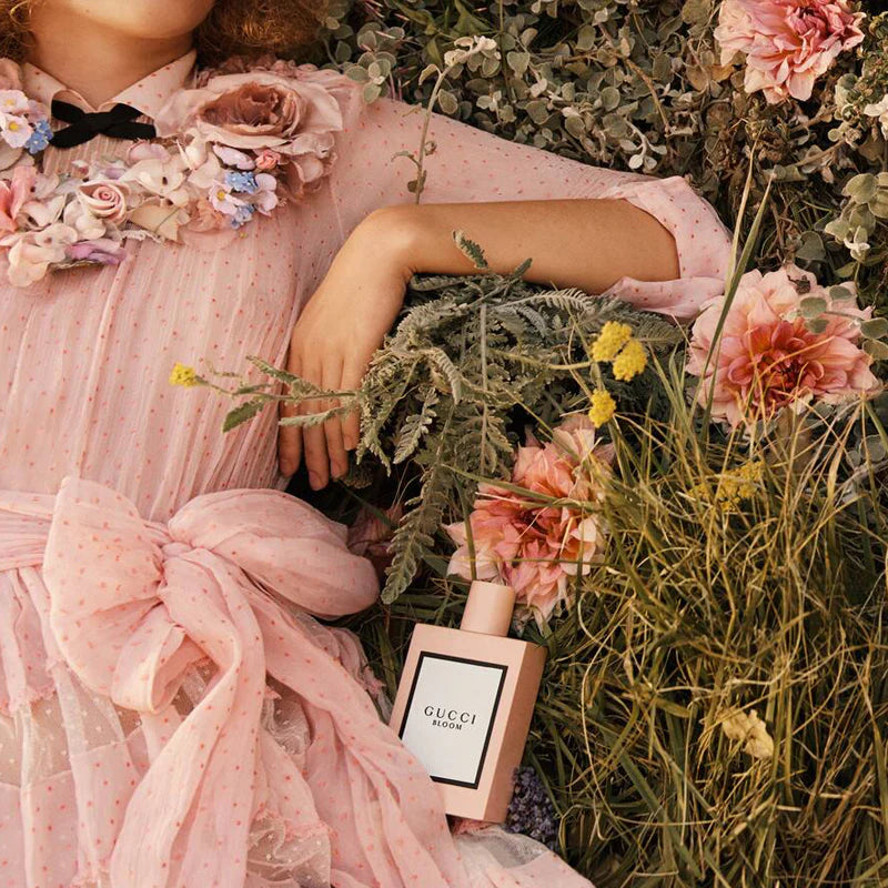 Woman in a pink dress with floral accessories lying among flowers and plants, Gucci perfume bottle visible.