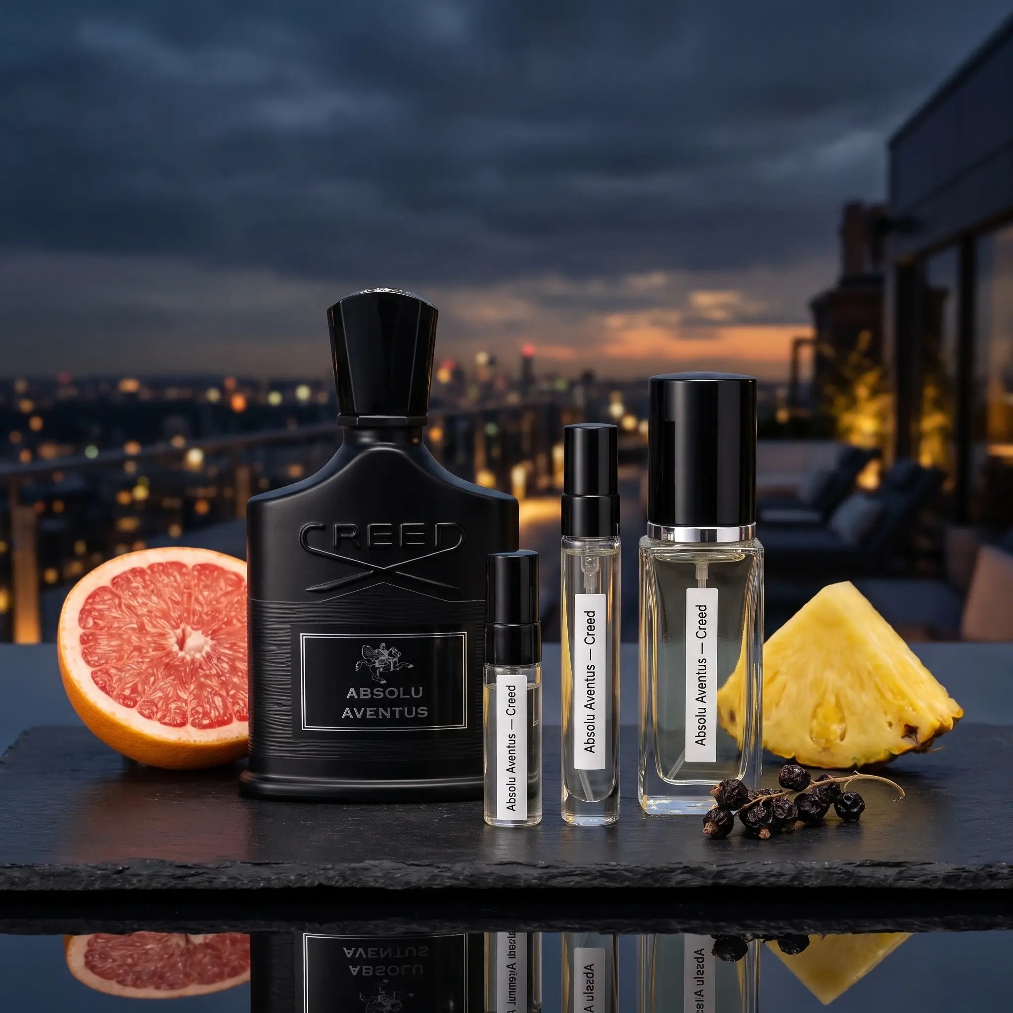 Creed Absolu Aventus perfume bottle displayed with two smaller sample vials on a dark tabletop, surrounded by a sliced grapefruit, pineapple, and peppercorns, with a city skyline at dusk in the background.