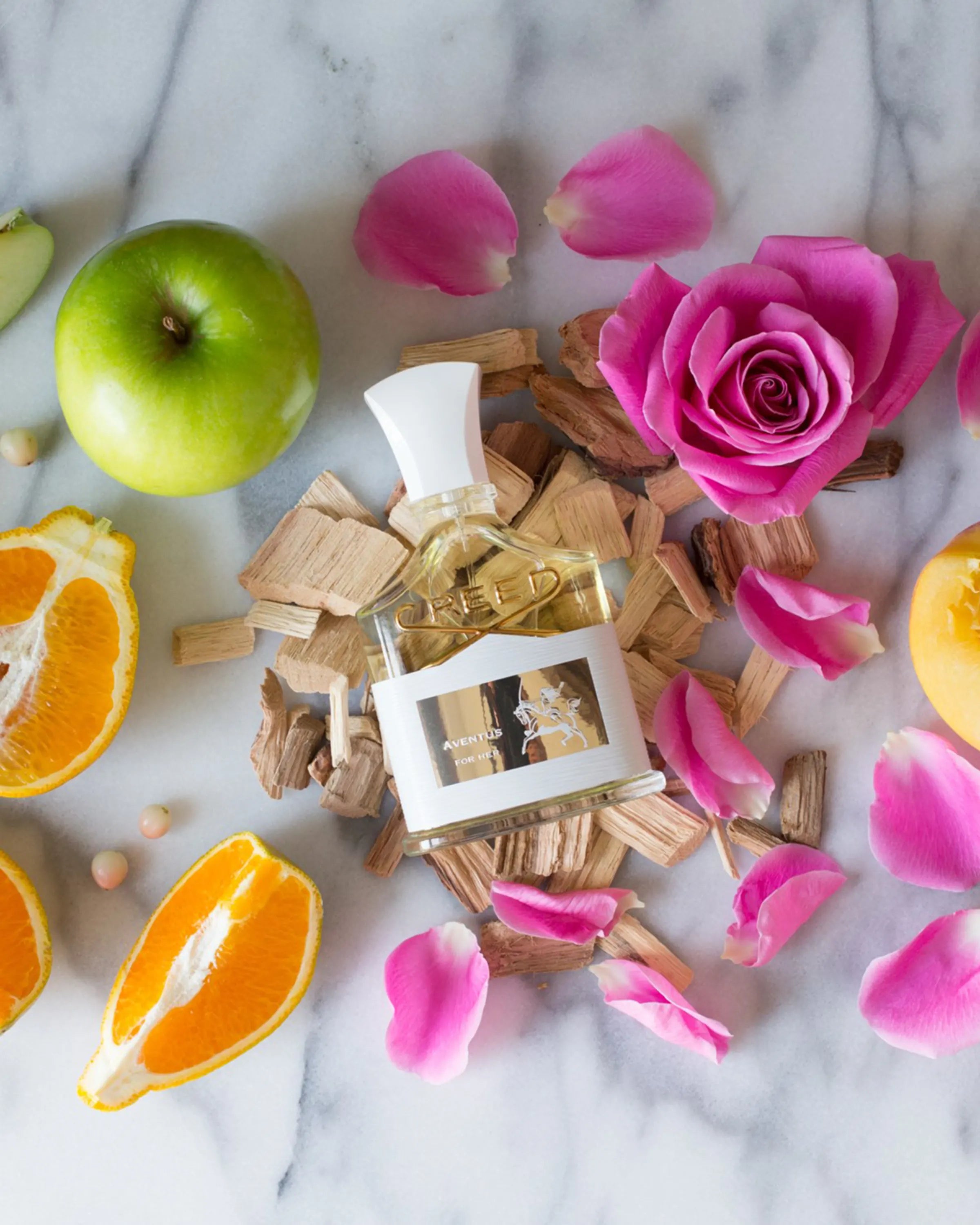 Perfume bottle surrounded by flowers, fruits, and wood on a marble surface