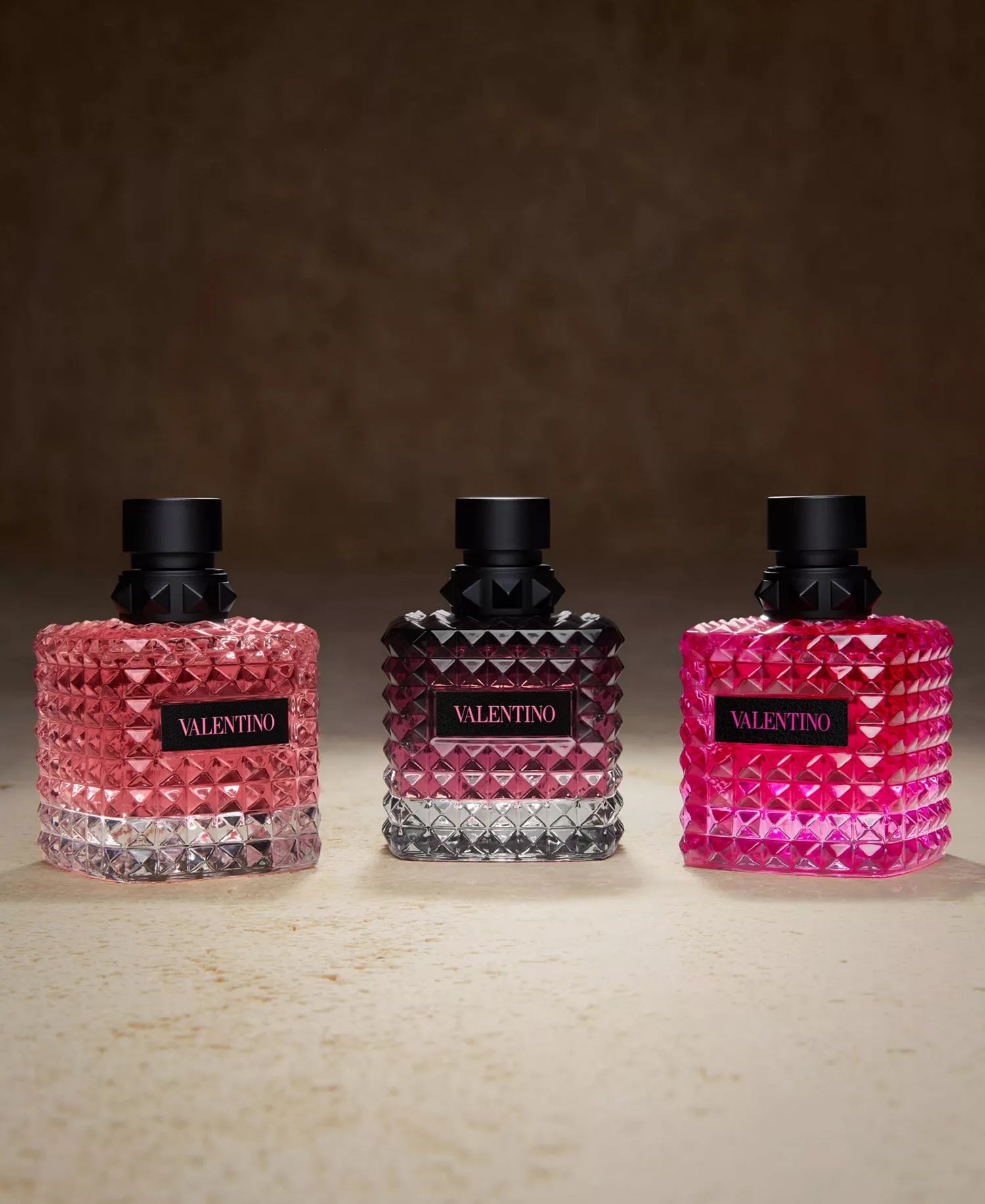 Three Valentina perfume bottles in pink, black, and red on a beige surface with a brown background.