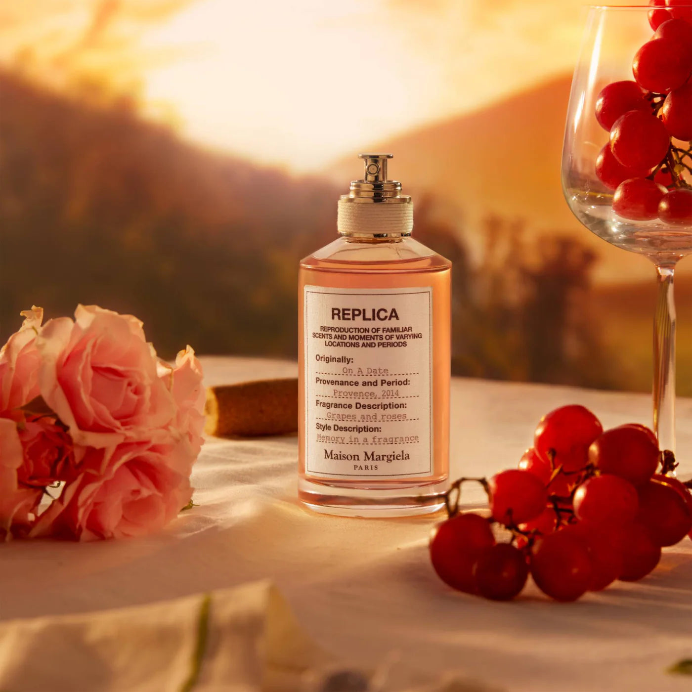Perfume bottle labeled 'Replica' with flowers and a glass of red berries on a table with a sunset background.