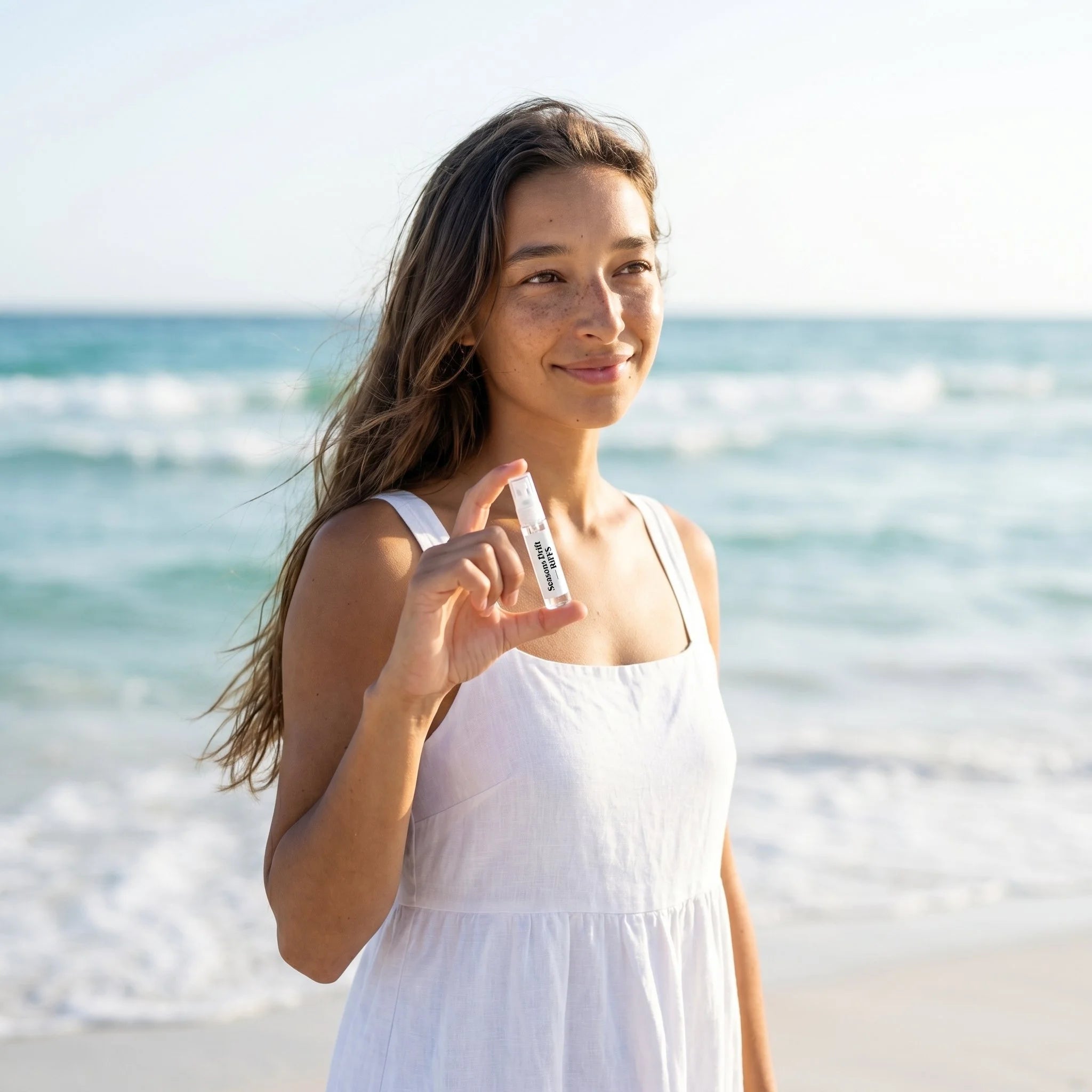 Woman holding RiiFFS Seasons Drift decant spray on the beach, smiling, summer coastal fragrance
